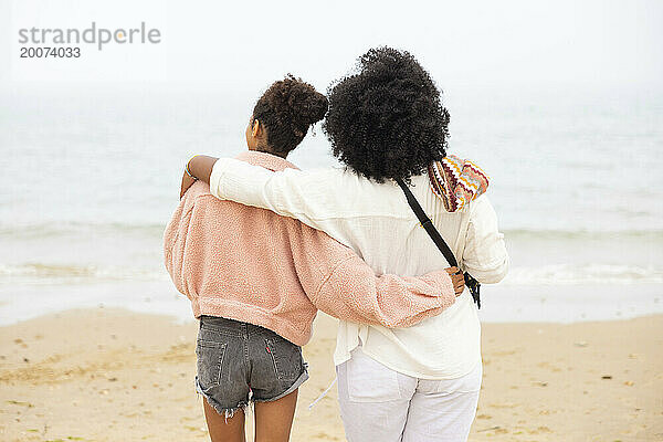 Zwei gemischtrassige Schwestern am Strand umarmen sich und blicken auf das Meer.