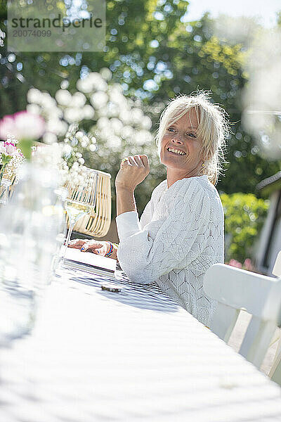 Attraktive blonde Mutter sitzt lachend und entspannt an ihrem Gartentisch in der Sonne. Verschwommener Garten und Blumen im Hintergrund. Glas Wein auf dem Tisch