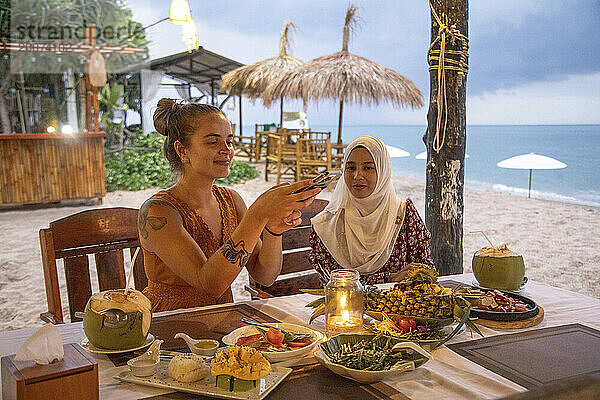 Zwei junge Freundinnen  eine davon trägt einen Hijab und isst in einem Strandrestaurant zu Abend und fotografiert das Essen