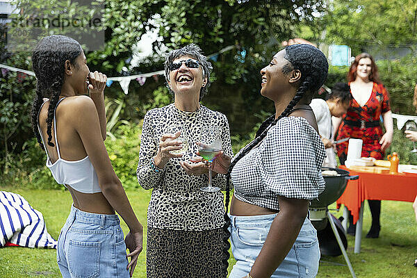 Ältere Frau lacht und hat Spaß mit zwei jungen Frauen/Enkeltöchtern/Freundinnen bei einer Grillparty im Sommergarten.