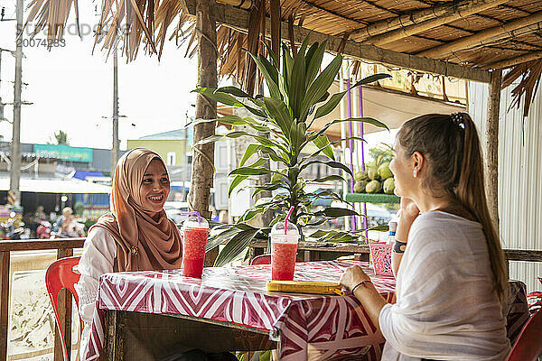 Zwei junge Frauen  eine davon trägt einen Hijab  sitzen zusammen in einem Café-Restaurant in einer ländlichen Stadt in Thailand