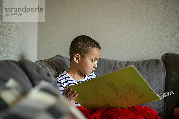 Kleiner Junge sitzt auf dem Sofa  isst gesunde Erdbeeren und liest ein Buch