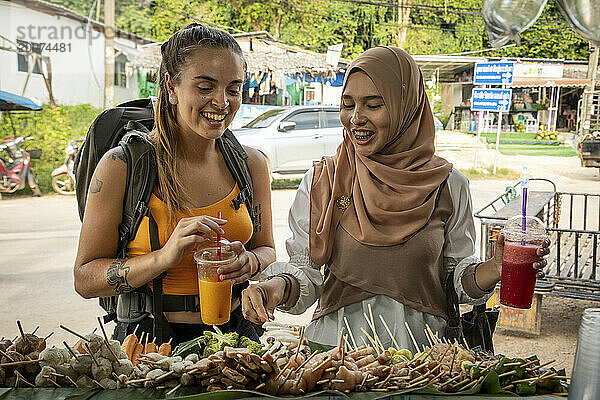 Zwei junge Frauen  eine davon trägt einen Hijab  lacht zusammen und kauft Straßenessen.