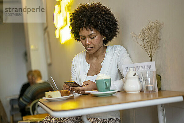 Attraktive Afrikanerin mit schönen Haaren sitzt mit ihrem Telefon in einem Café