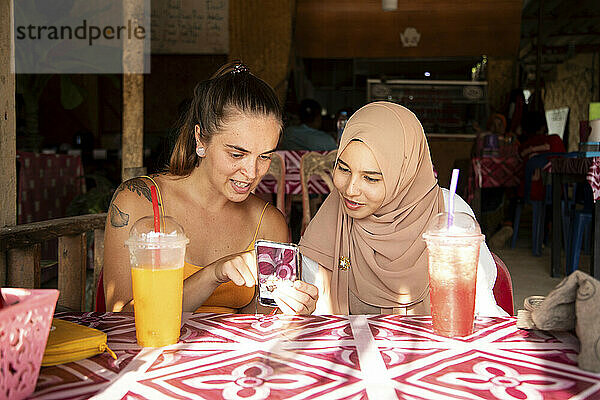 Zwei junge Freundinnen  eine davon trägt einen Hijab  sitzen zusammen in einem Restaurant  spielen mit ihren Handys und zeigen sich gegenseitig Fotos