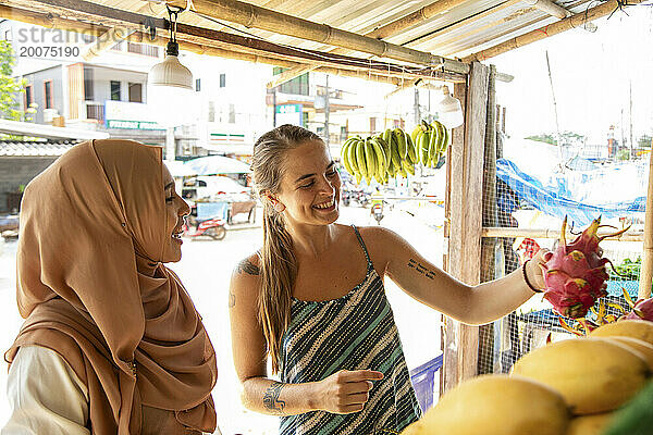 Zwei junge Frauen  eine davon trägt einen Hijab und kauft Drachenfrüchte in einer ländlichen Stadt in Thailand