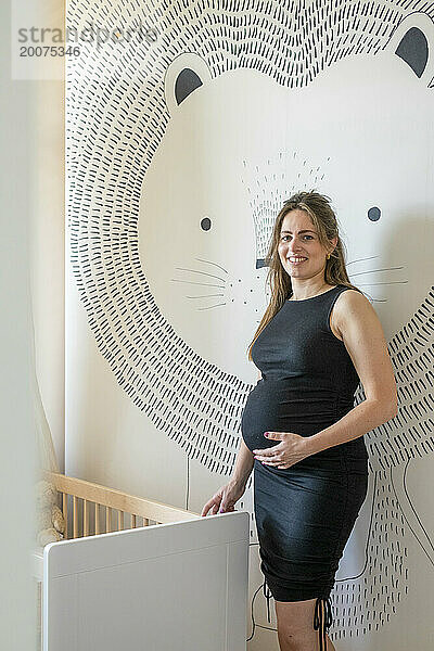 Fröhliches Porträt einer schwangeren Mutter  die neben ihrem Kinderbett steht