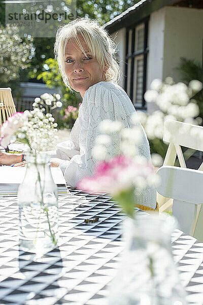 Attraktive blonde Mutter sitzt lachend und entspannt an ihrem Gartentisch in der Sonne. Verschwommener Garten und Blumen im Hintergrund