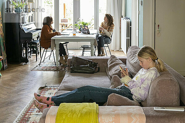 Junger Teenager entspannt sich auf dem Sofa und recherchiert mit ihrem Telefon. Im Hintergrund sitzen Großmutter und Mutter bei einer Tasse Tee