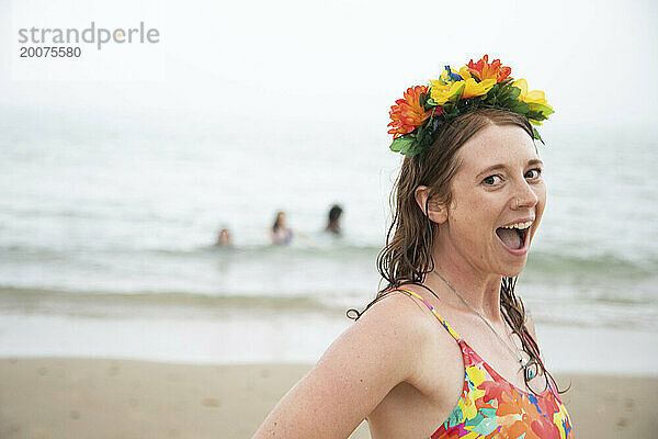 Eine junge weiße Frau lächelt begeistert in die Kamera und vergnügt sich mit Blumen im Haar am Strand