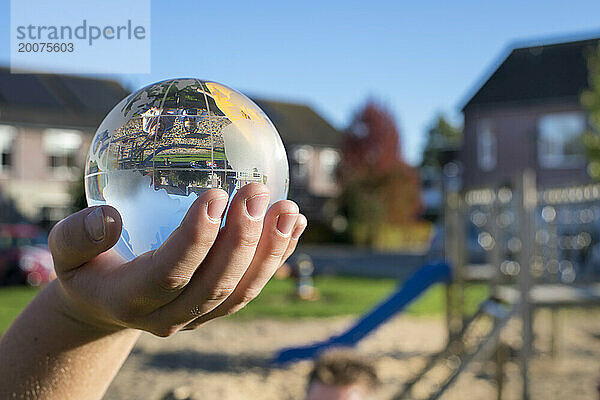 Kind hält eine Glaskugel in einem öffentlichen Park  reflektierendes Licht und klares Glas