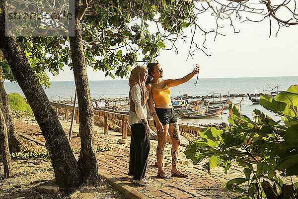 Zwei junge Freundinnen  von denen eine einen Hijab trägt  gehen den Küstenweg im Süden Thailands entlang und haben angehalten  um ein Selfie zu machen.