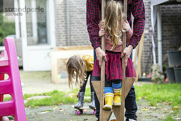 Vater hilft seiner Tochter beim Balancieren auf Stelzen im Garten und hat Spaß. Ihre Schwester im Hintergrund versucht Skateboard zu fahren