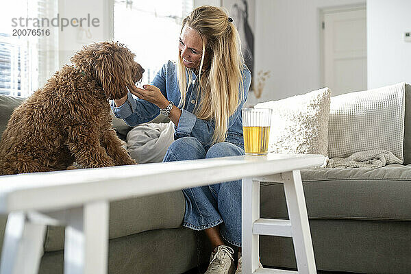 Attraktive blonde Frau sitzt auf ihrem Sofa und spricht mit ihrem Hund  der ihm Zuneigung schenkt
