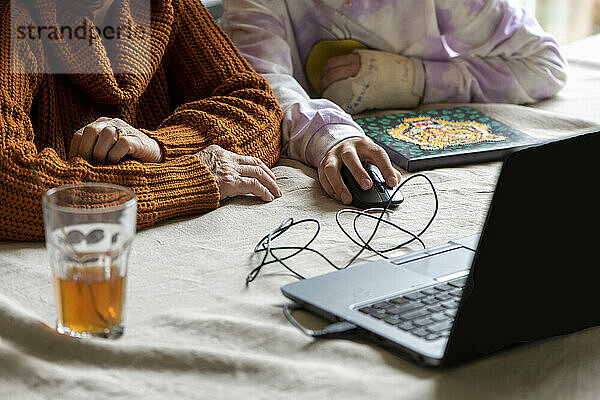 Junge Enkelin im Teenageralter hilft einer älteren Frau bei der Recherche und dem Ausfüllen von Formularen mit ihrem Laptop und Telefon. Die Familie um sie herum unterstützt sie