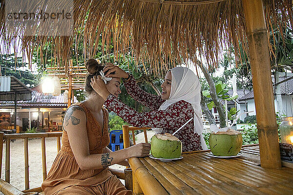 Zwei junge Freundinnen  eine davon trägt einen Hijab in einem Strandrestaurant  sie steckt ihrer Freundin eine Blume ins Haar.