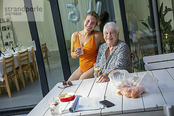 Enkelin und ältere Frau sitzen um ein Kabel herum und feiern ihren Geburtstag mit Wein und Essen. Luftballons im Hintergrund