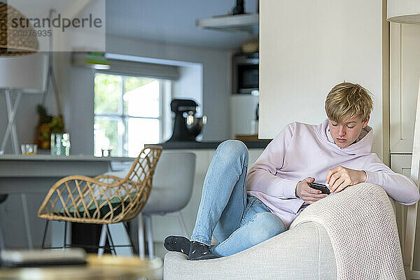 Junger Mann sitzt auf dem Sofa in einem sauberen  modernen Zuhause und surft auf seinem Telefon