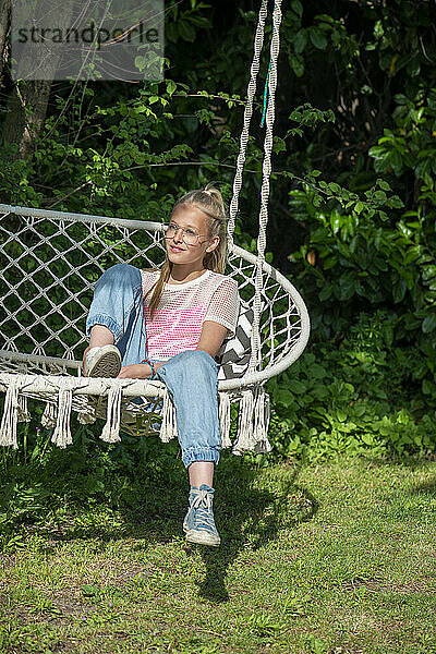 Blondes Teenager-Mädchen sitzt zu Hause auf einem Stuhl und genießt die Sonne und blickt in Frieden und glücklich in die Kamera