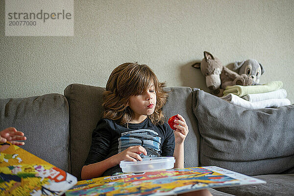 Kleiner Junge sitzt auf dem Sofa  isst gesunde Erdbeeren und liest ein Buch