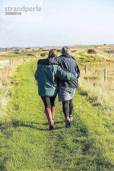 Mutter und erwachsene Tochter spazieren zusammen am Fluss Ore  Suffolk  England  UK von hinten umarmend