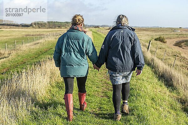 Mutter und erwachsene Tochter spazieren zusammen am Fluss Ore  Suffolk  England  UK von hinten und halten sich an den Händen