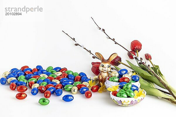 Eine Ansammlung von bunten Schokoladeneiern und einem Osterhasen neben Tulpen und Palmkätzchen  weißer Hintergr