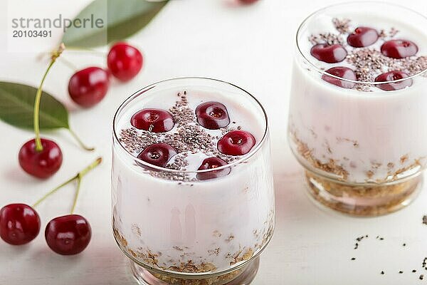 Joghurt mit Kirschen  Chiasamen und Müsli im Glas mit Holzlöffel auf weißem Holzhintergrund. Seitenansicht  Nahaufnahme  selektiver Fokus