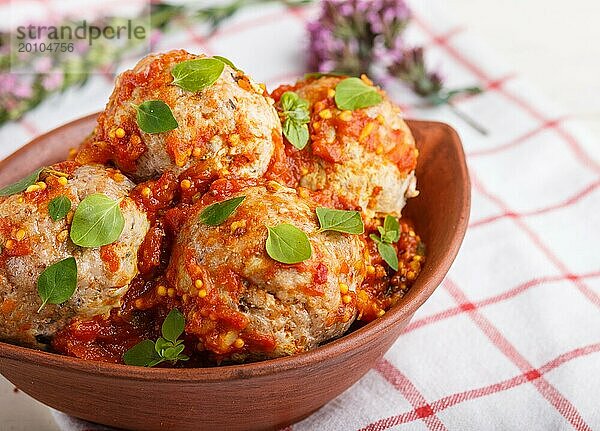 Schweinefleischbällchen mit Tomatensoße  Oreganoblättern  Gewürzen und Kräutern in einer Tonschale auf weißem Holzhintergrund mit Leinentextil. Seitenansicht  Nahaufnahme  selektiver Fokus