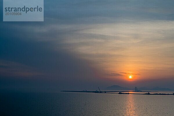 Baukähne und Kräne als Silhouetten gegen den bewölkten Himmel und das Meerwasser bei Sonnenuntergang in Südkorea