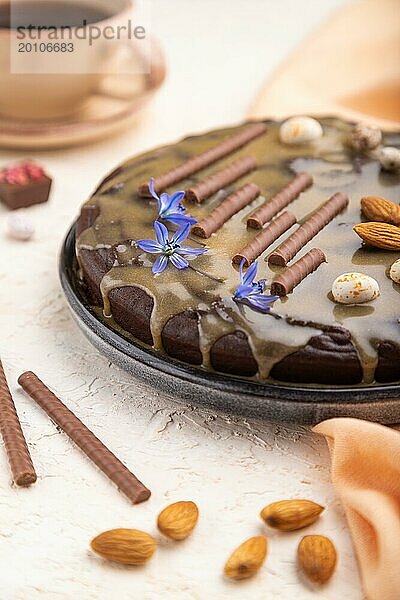 Hausgemachter Schokoladen Brownie Kuchen mit Karamellcreme und Mandeln mit einer Tasse Kaffee auf einem weißen Betonhintergrund und orangefarbenem Textil. Seitenansicht  Nahaufnahme  selektiver Fokus