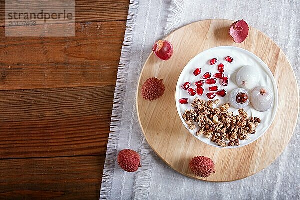 Griechischer Joghurt mit Litschi  Granatapfelkernen und Granola in einem weißen Teller auf einem braunen Holzhintergrund  Draufsicht  Flat Lay  Copy Space  selektiver Fokus