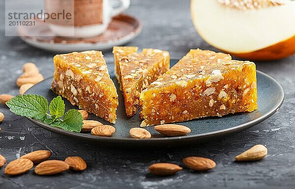 Traditionelle türkische Süßigkeit Cezerye aus karamellisierter Melone  gerösteten Walnüssen  Haselnüssen  Cashew  Pistazien in blauem Keramikteller und einer Tasse Kaffee auf schwarzem Betonhintergrund. Seitenansicht  Nahaufnahme  selektiver Fokus