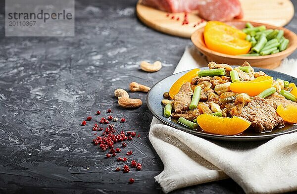 Gebratenes Schweinefleisch mit Pfirsichen  Cashewnüssen und grünen Bohnen auf einem schwarzen Betonhintergrund. Seitenansicht  Kopierraum  selektiver Fokus  chinesische Küche. Leinenstoff