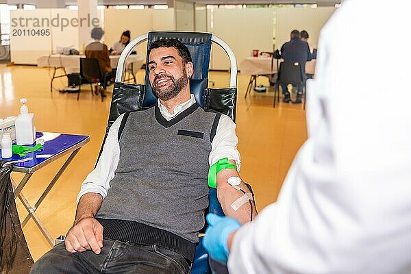 Ein Mann sitzt lächelnd auf einem Stuhl neben einem nicht erkennbaren Arzt in einem Blutspendepavillon