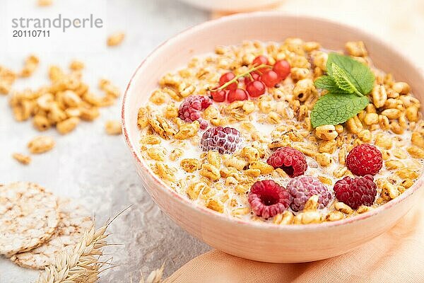 Weizenflockenbrei mit Milch  Himbeeren und Johannisbeeren in einer Keramikschale auf grauem Betonhintergrund und orangefarbenem Leinentextil. Seitenansicht  Nahaufnahme  selektiver Fokus