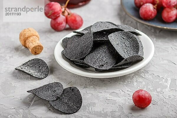 Schwarze Kartoffelchips mit Holzkohle  Balsamico Essig im Glas  rote Trauben auf einem blauen Keramikteller auf einem grauen Betonhintergrund. Seitenansicht  Nahaufnahme  selektiver Fokus