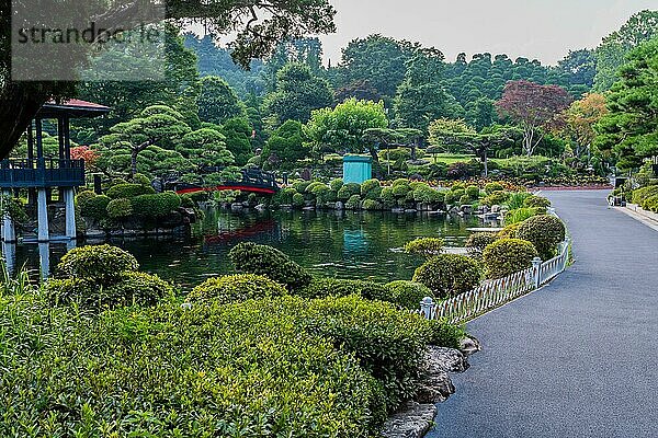 Gepflasterter Weg neben einem Koi Teich mit einem Steg  der zu einem Pavillon in einem gepflegten öffentlichen Park in Südkorea führt