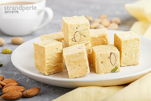 Traditionelle indische Süßigkeit soan papdi in weißem Teller mit Mandel  Pistache und einer Tasse Kaffee auf einem grauen Betonhintergrund mit gelbem Textil. Seitenansicht  Nahaufnahme  selektiver Fokus