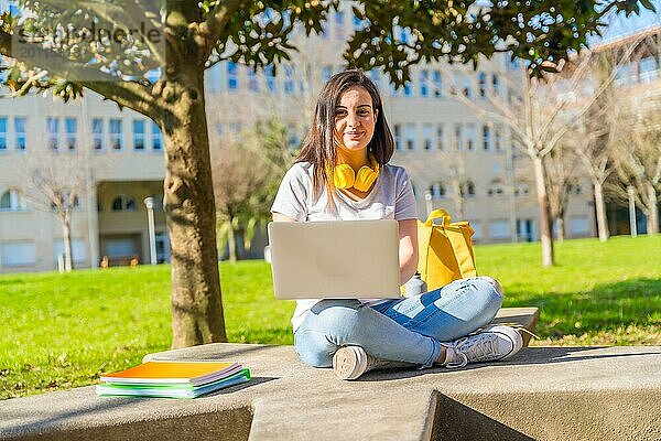 Porträt eines fröhlichen Schülers  der mit seinem Laptop auf dem Boden vor dem Campus sitzt