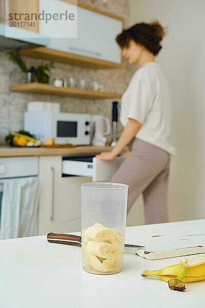 Eine Schüssel mit einem Stabmixer und geschnittenen Bananen auf dem Küchentisch. Im Hintergrund  unscharf  holt eine Frau Besteck aus dem Schrank