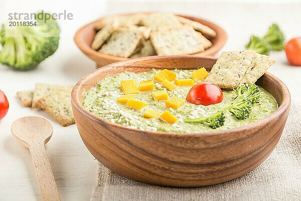 Grüne Brokkolicremesuppe mit Crackern und Käse in Holzschale auf weißem Holzuntergrund und Leinenserviette. Seitenansicht  Nahaufnahme  selektiver Fokus