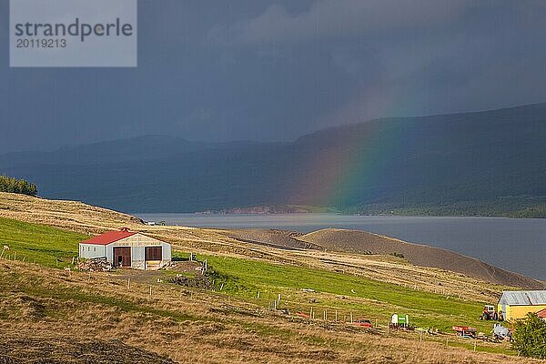 Regenbogen  der sich über einem breiten Fluss in einem Tal mit einem kleinen weiß roten Bauernhof auf Island ausbreitet