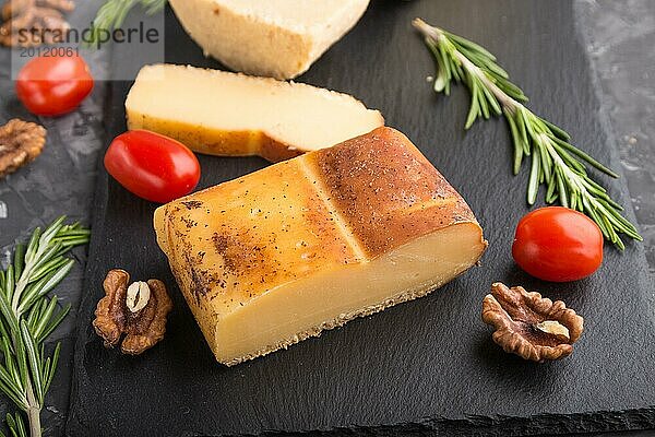 Geräucherter Käse und verschiedene Käsesorten mit Rosmarin und Tomaten auf einer schwarzen Schieferplatte auf einem schwarzen Betonhintergrund. Seitenansicht  Nahaufnahme  selektiver Fokus