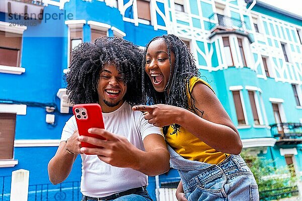 Afrikanische Freunde  die mit überraschtem Gesichtsausdruck auf ein Handy zeigen  das neben blauen bunten Häusern in der Stadt steht