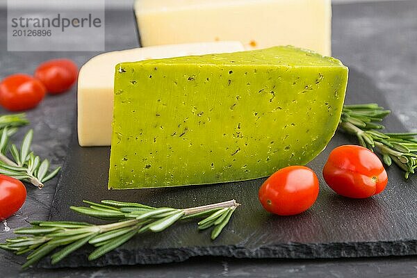 Grüner Basilikumkäse und verschiedene Käsesorten mit Rosmarin und Tomaten auf einer schwarzen Schiefertafel auf einem schwarzen Betonhintergrund. Seitenansicht  Nahaufnahme  selektiver Fokus