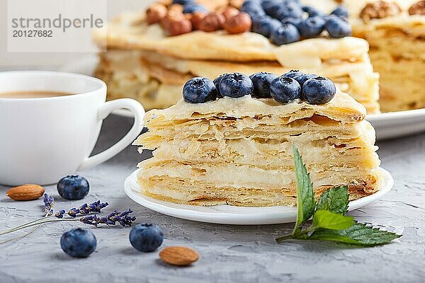 Hausgemachte geschichteten Napoleon Kuchen mit Milch Creme. Dekoriert mit Heidelbeere  Mandeln  Walnüsse  Haselnüsse  Minze auf einem grauen Beton Hintergrund und Tasse Kaffee. Seitenansicht  Nahaufnahme  selektiver Fokus