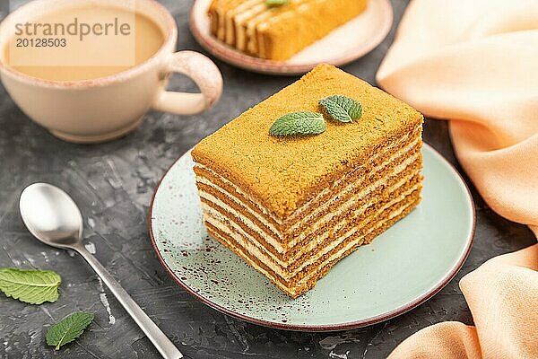 Hausgemachter Honigkuchen mit Milchcreme und Minze mit einer Tasse Kaffee auf einem schwarzen Betonhintergrund und orangefarbenem Textil. Seitenansicht  Nahaufnahme  selektiver Fokus