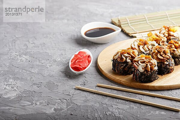 Japanische Maki Sushi Rollen mit Frischkäse  Essstäbchen  Sojasauce und mariniertem Ingwer auf einem Holzbrett auf grauem Betonhintergrund. Seitenansicht  Kopierraum  selektiver Fokus