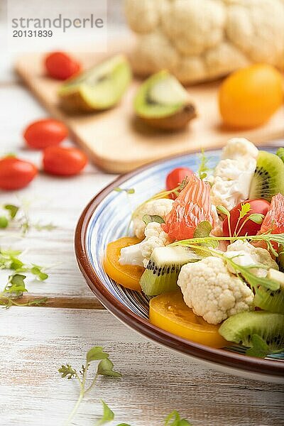 Vegetarischer Salat aus Blumenkohl  Kiwi  Tomaten  Mikrogrünsprossen auf weißem Holzhintergrund und blauem Leinentuch. Seitenansicht  Nahaufnahme  selektiver Fokus